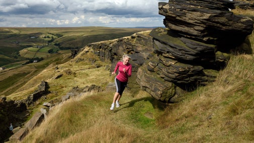 A women runs past a rocky outcrop on a hillside, with a landscape of rolling hills behind her